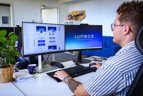 Un homme travaille sur deux écrans d'ordinateur dans un bureau moderne avec le logo Luneos.