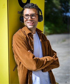 Homme souriant avec écouteurs, devant un panneau jaune "L'agence